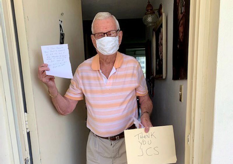 Older male wearing a face mask holding a thank you JCS sign as he stands in front of his doorway