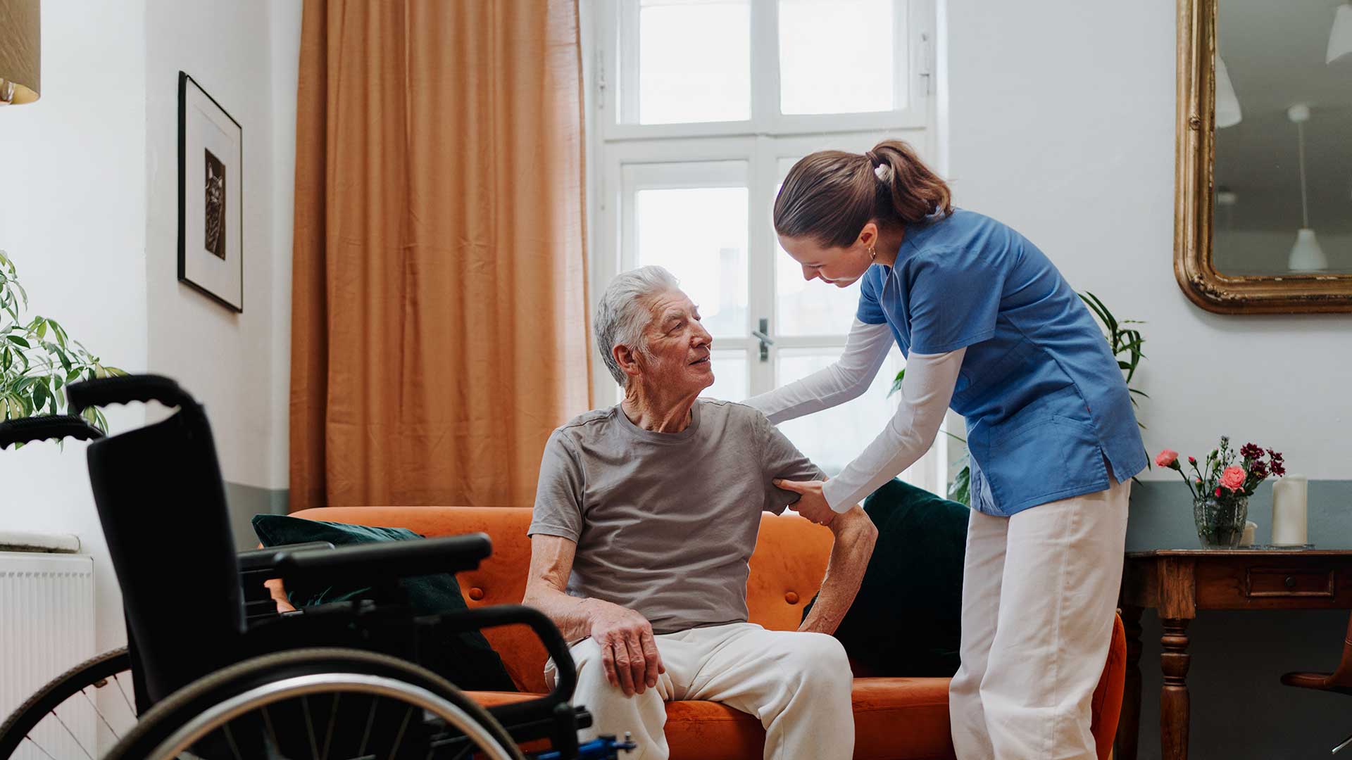 caretaker helps an elderly man off the couch to his wheelchair